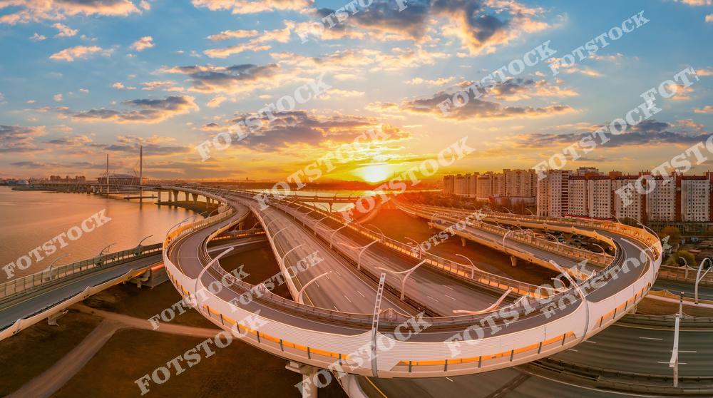 На фотографии изображен Западный скоростной диаметр (ЗСД) в Санкт-Петербурге, освещенный лучами закатного солнца. Изящные линии магистрали плавно изгибаются, соединяя районы города. Золотые оттенки неба и облаков подчеркивают красоту инженерного сооружения, а современные жилые дома завершают композицию. Этот снимок демонстрирует гармонию между природой, архитектурой и транспортной инфраструктурой.